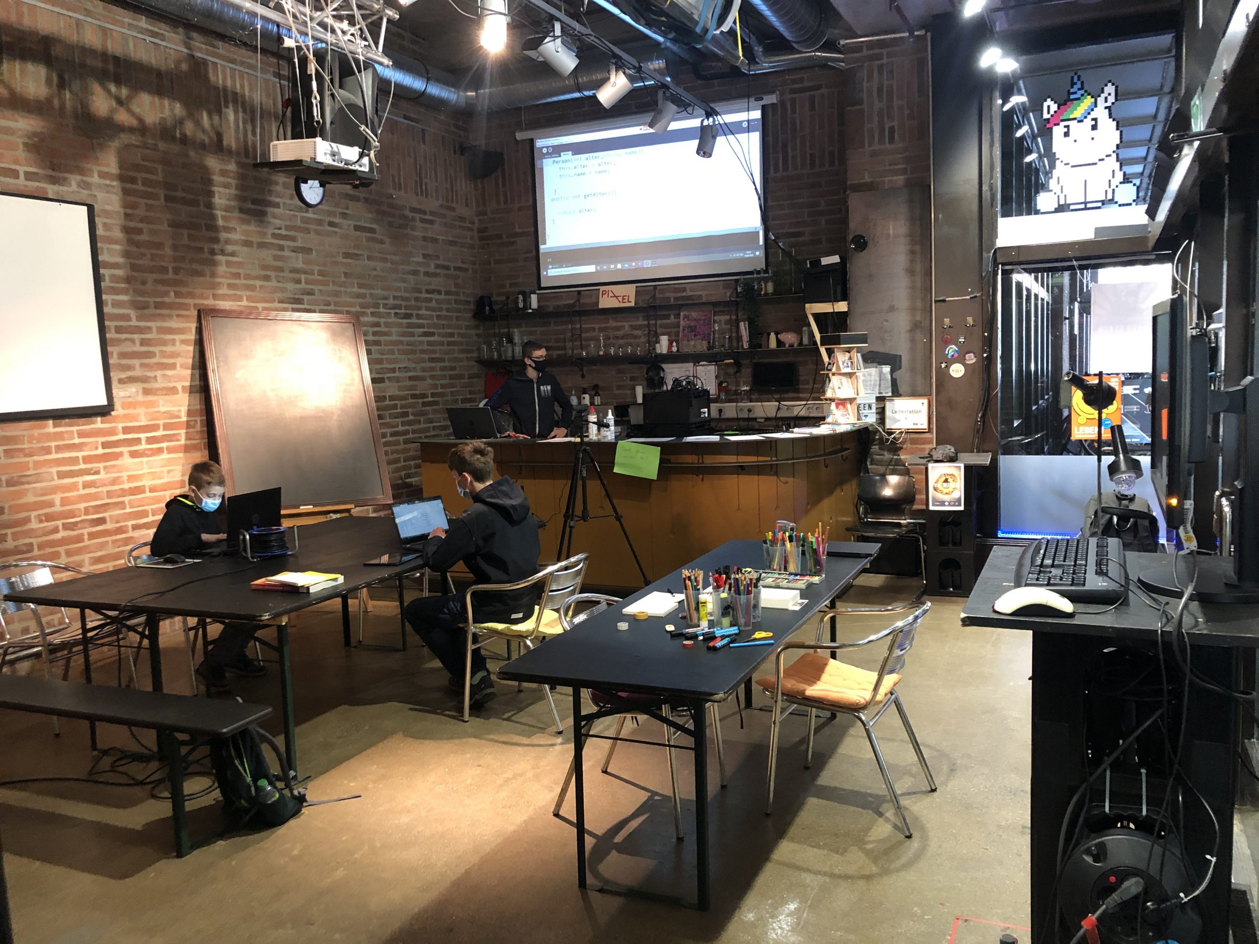 Programmierkurs Java/Processing. In einem Raum sitzen zwei jüngere Kinder an zwei Laptops. Es ist eine Leinwand aktiv, jemand steht davor und erklärt an ihr etwas über Codierung.