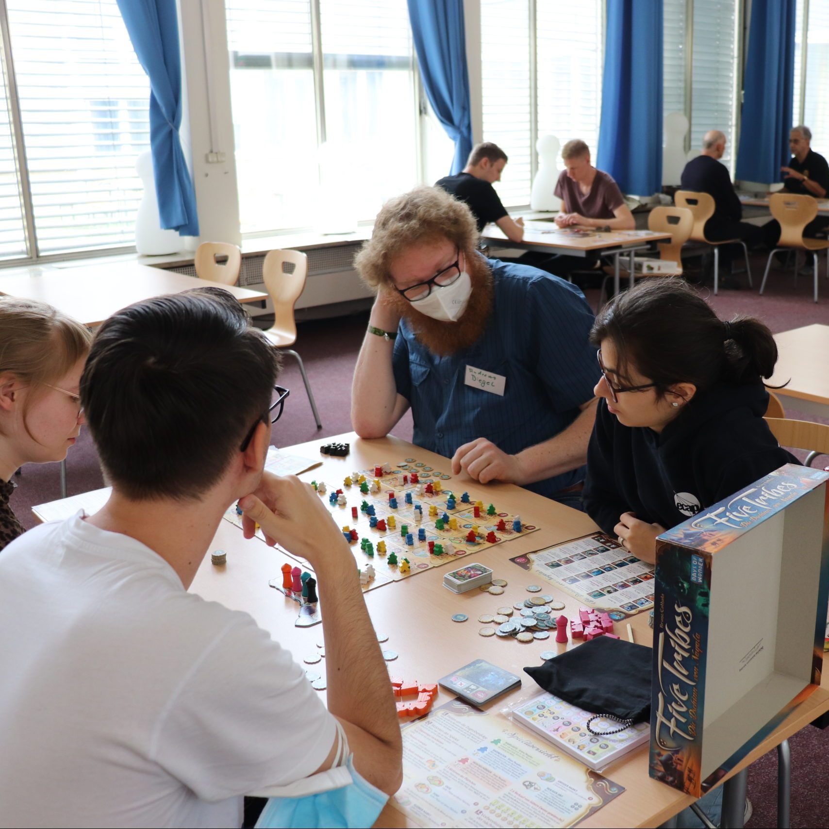 In einem hellen Saal sind verschiedene Tische mit Stühlen platziert. Im Hintergrund sieht man zwei junge Menschen gemeinsam am Tisch ein Spiel spielen, etwas dahinter auch zwei Ältere an einem anderen. Im Vordergund sitzen vier Menschen, ebenfalls an einem Tisch und spielen das Brettspiel Five Tribes. Die vier Beteiligten sehen konzentriert aufs Spielbrett mit vielen bunten Spielfiguren.