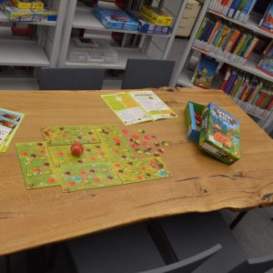 Ein großer Holztisch in einer Bibliothek, auf ihm ist das Brettspiel Speedy Roll aufgebaut, mit verschiedenen Spielfeldern, drei Anleitungen, einem kleinen Ball und der Verpackung