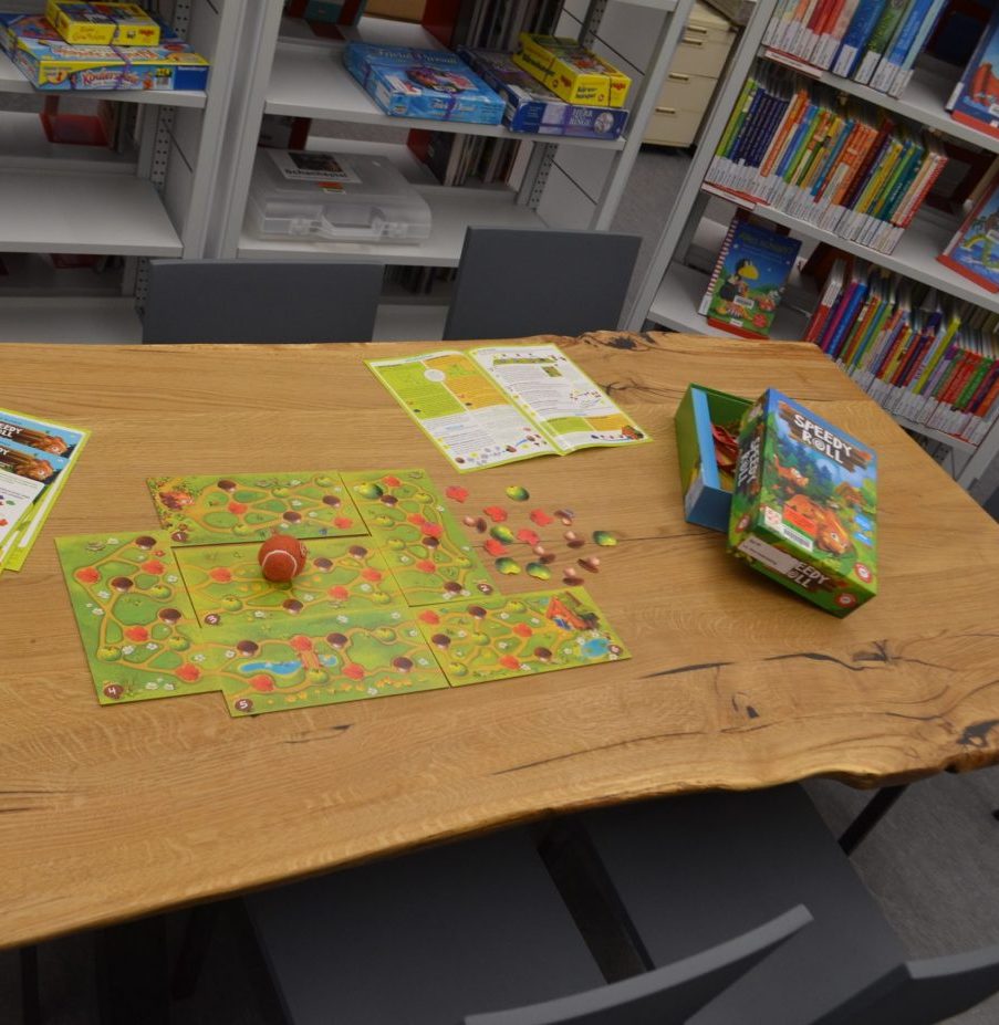 Ein großer Holztisch in einer Bibliothek, auf ihm ist das Brettspiel Speedy Roll aufgebaut, mit verschiedenen Spielfeldern, drei Anleitungen, einem kleinen Ball und der Verpackung
