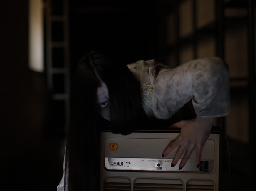 Cosplayerin verkleidet als das Mädchen aus dem Film "The Ring", blickt in die Kamera und klettert über einen alten Fernseher. Der Hintergrund ist sehr dunkel, das Mädchen in weißen, dreckigen Klamotten und mit weißem Make-Up im Gesicht, sticht heraus. Es handelt sich um ein Cosplayshooting.